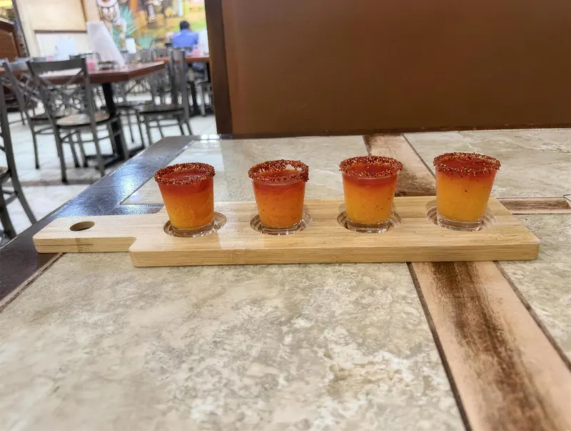 Four chilli-rimmed tequila shots lined up on a wooden paddle board