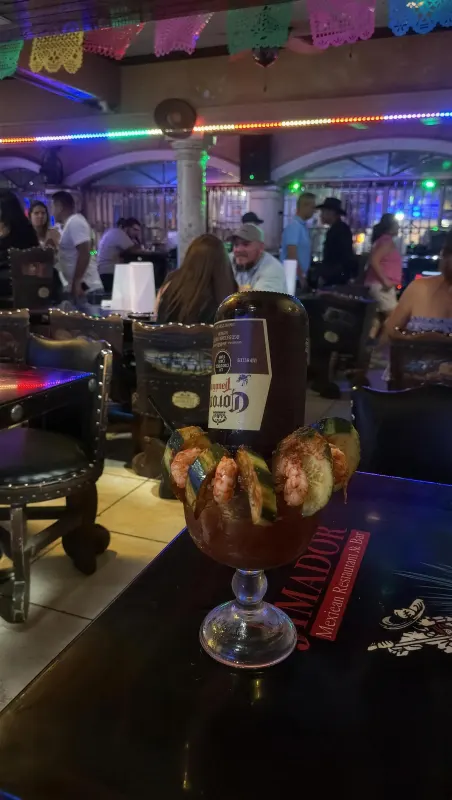 A michelada garnished with shrimp and cucumber on a table at night, the lit dining room and arched entrance glowing behind it under strings of papel picado