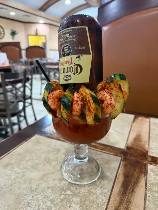 Michelada served in a tall glass, rimmed with chilli and garnished with shrimp and cucumber, a Corona Familiar bottle inverted in it